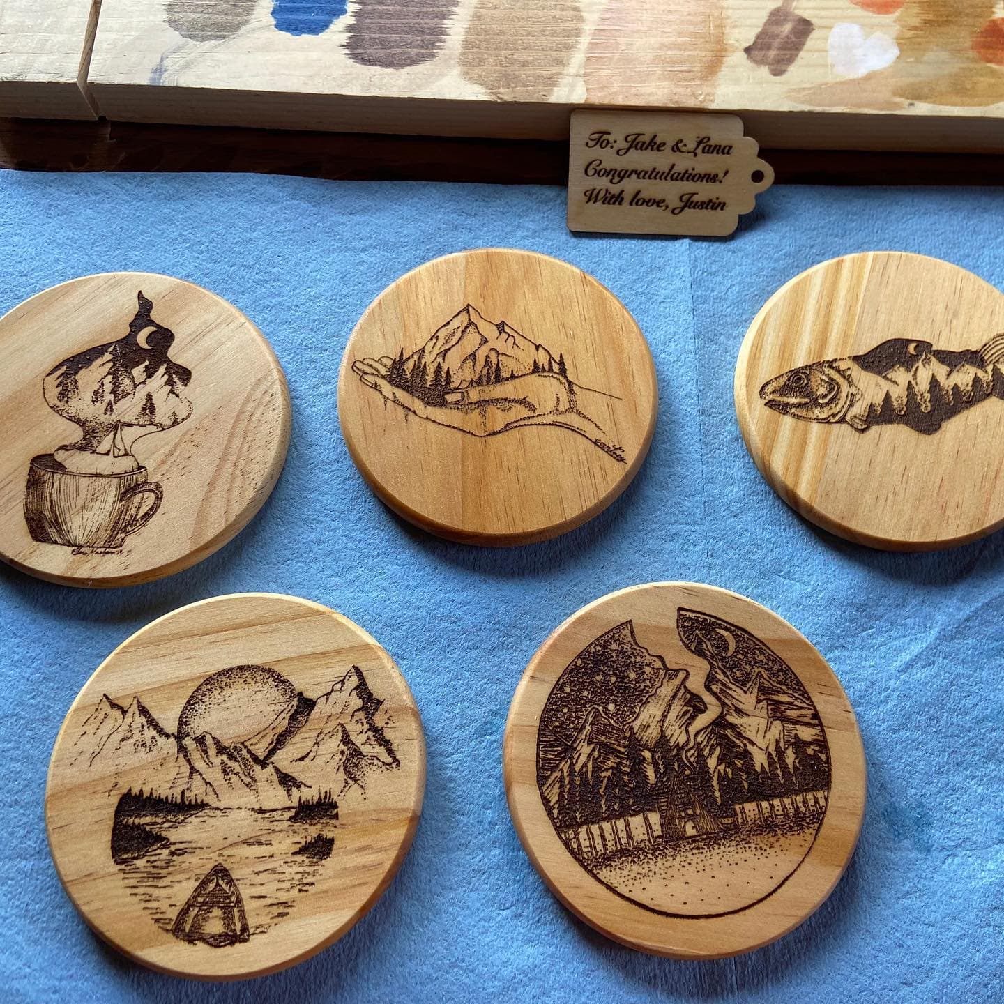 Five wooden coasters laser-engraved with mountain, fish, and landscape scenes, with a gift tag